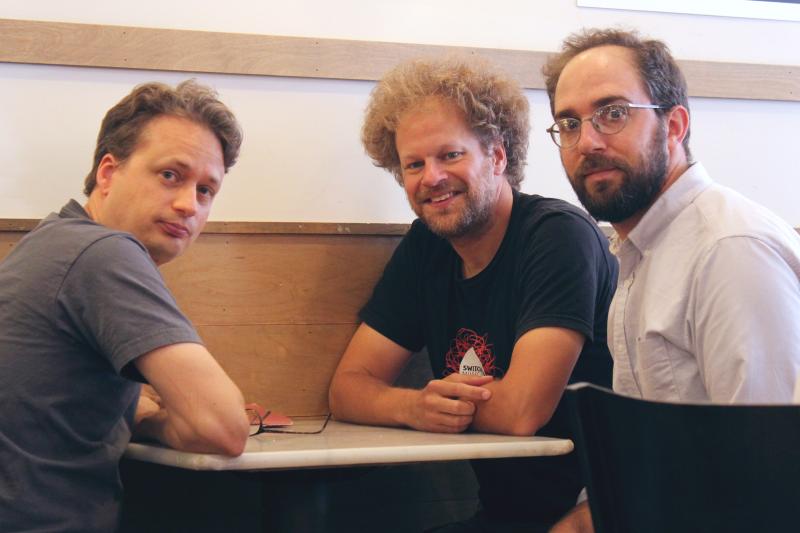 The three founders of Experiments in Opera Three men sitting at a table looking at camera