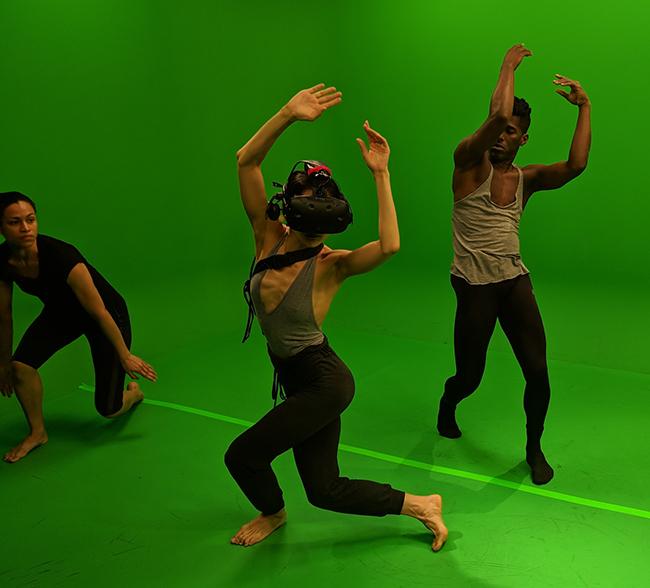 A dancer wearing virtual reality headgear performs with other dancers in a green room