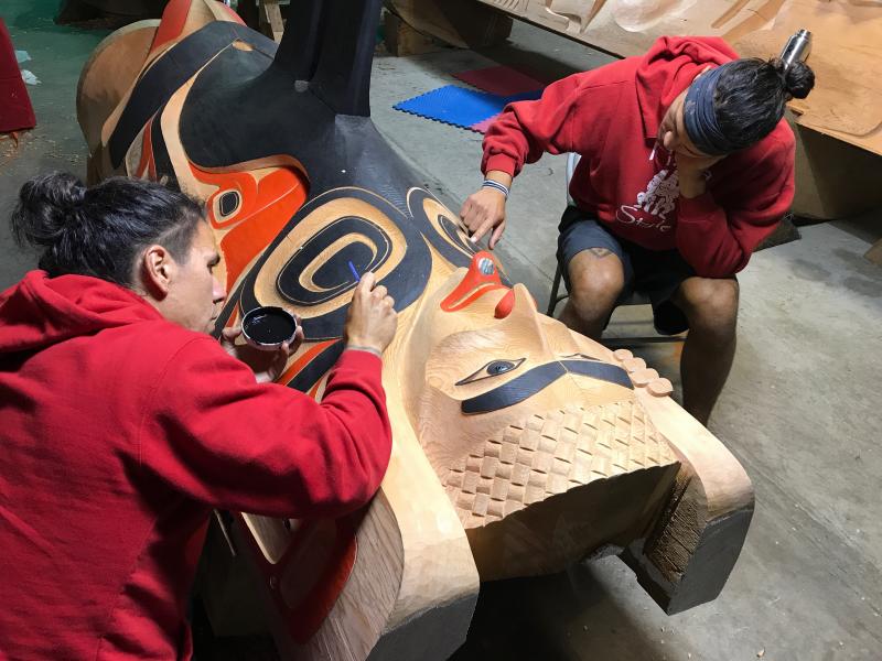 Two men wearing red carve a totem pole