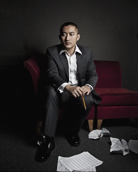 Huang Ruo sits on a red velvet couch holding a pencil. Music composition paper is strewn at his feet