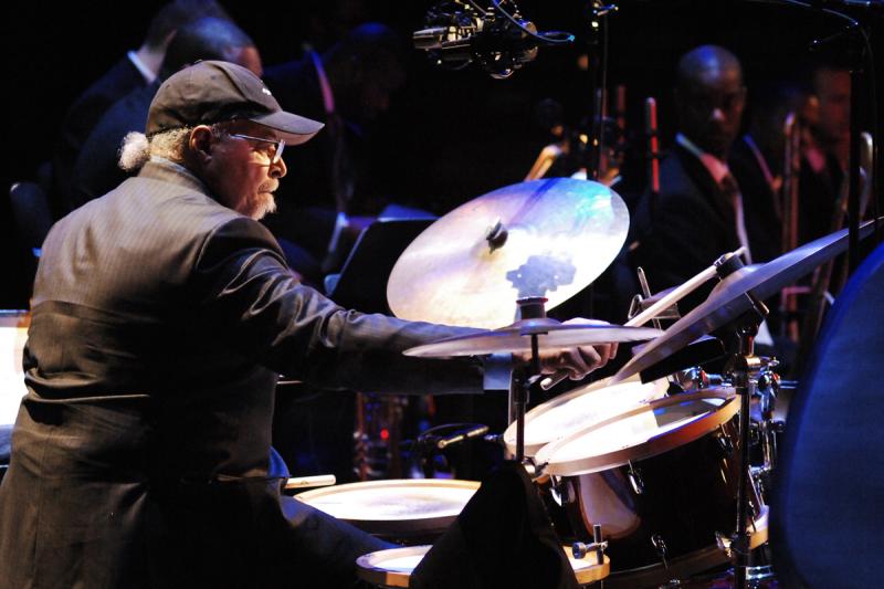 A man wearing a black baseball hat plays the drums.