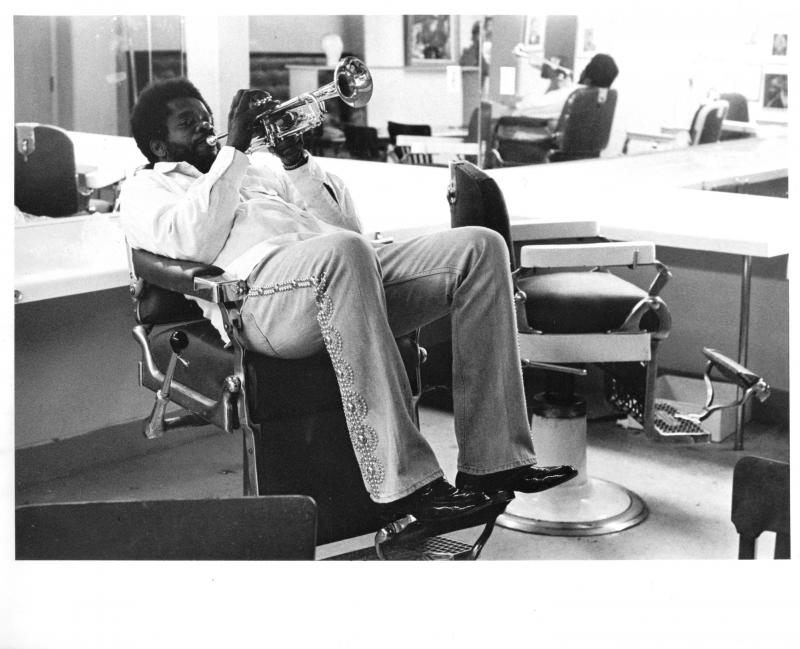 Man sitting in barber chair blowing a trumpet. 