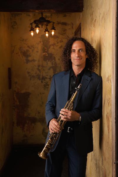 a full-shot of musician Kenny G wearing a dark blue suit and holding a soprano saxophone