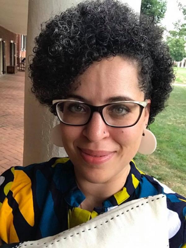 Kiki Petrosino Headshot of woman with curly short black hair, big black glasses and large dangly earrings
