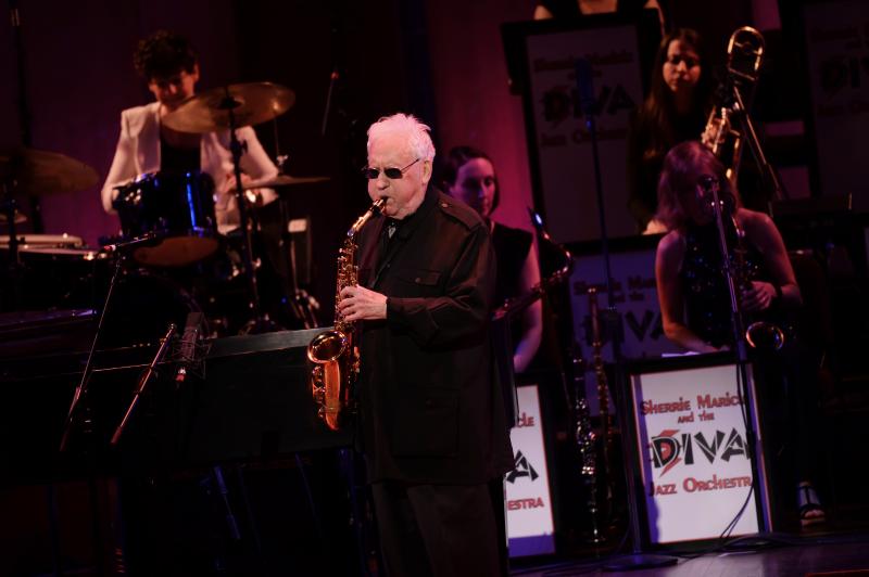 A man in all black wears sunglasses and plays the saxophone on a stage with other musicians behind him.