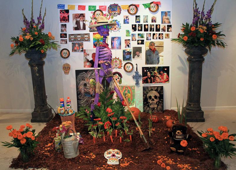 An altar for a Day of the Dead celebration with a purple skeleton, orange flowers, and a poster with photos on it.