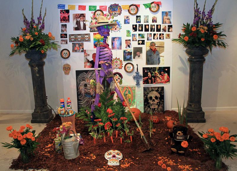 An altar for a Day of the Dead celebration with a purple skeleton, orange flowers, and a poster with photos on it. 