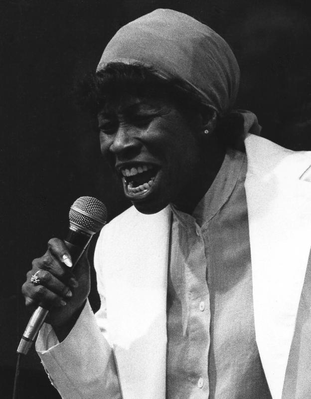 Woman with kerchief on her head singing into microphone. 