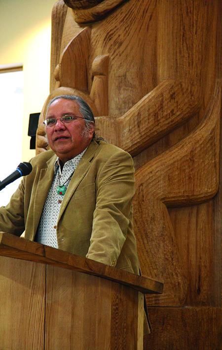 Native-American man at podium in front of large wooden carving. 