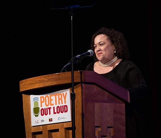 Pam Breaux at POL A woman speaks at a podium with a Poetry Out Loud banner hanging from it
