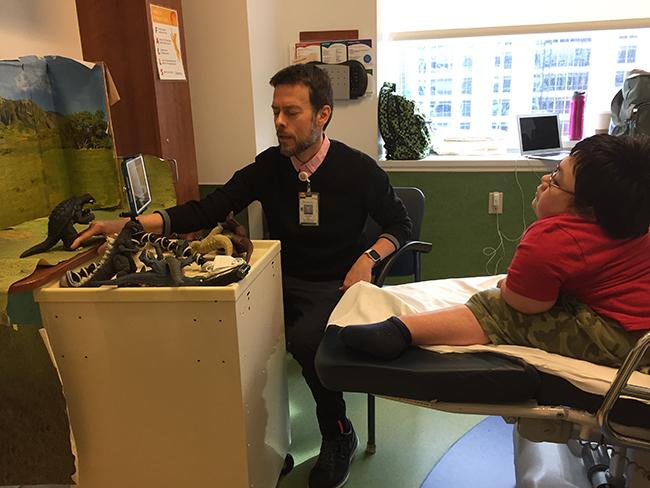 A boy in a hospital bed talks to a man working with Godzilla action figures as they direct a stop-motion film about Godzilla together