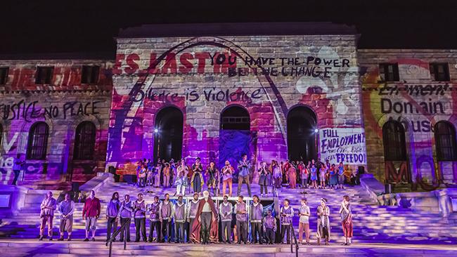 Performers line the steps of s stone building with art projections on it at night