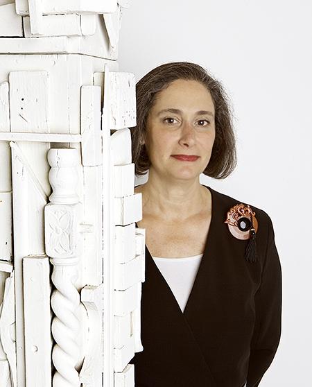 Woman wearing a black suit with large red brooch standing next to a white sculpture.