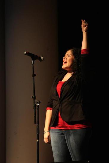 an indigenous American woman stands in front of a microphone one arm thrust in the air