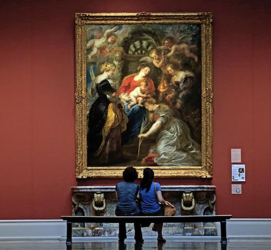 Two young women sit in front of a painting by Rubens of a group of women surrounded by putti