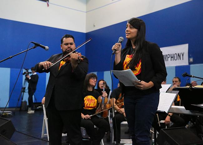 A man playing a violin stands next to a woman speaking into a microphone