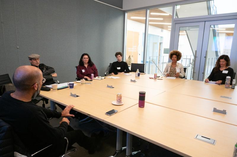 Six people, engaged in conversation, sit roundtable in a room with glass windows to the right. On the table, are an assortment of cups.