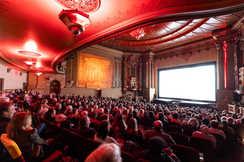 A large audience is seated inside of an auditorium, facing a large screen.