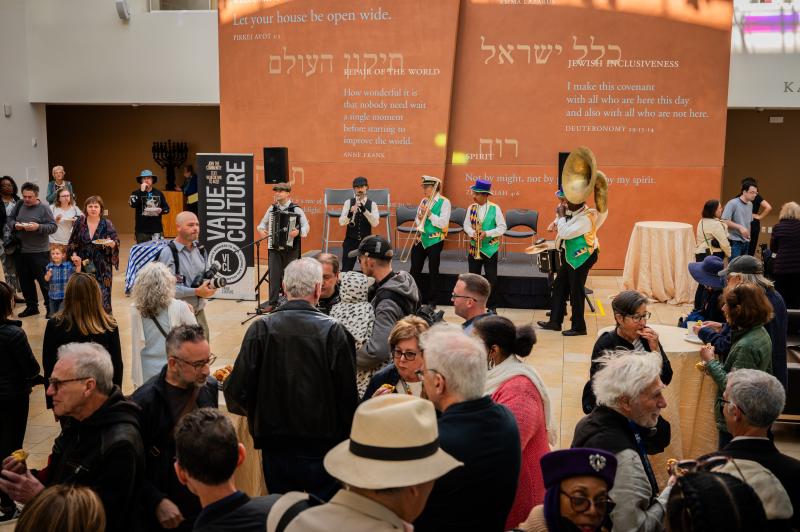 A large group of people are standing in a room watching live music (from instruments such as the trombone, accordion, and trumpet) being played.
