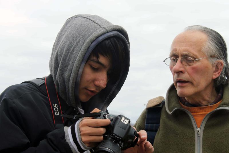 A teen wearing a hooded sweatshirt shows an image on his camera to an older man in a green fleece and glasses