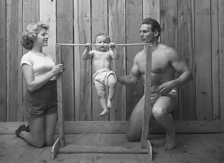 A mother and father stand on either side of a baby doing a pull-up 