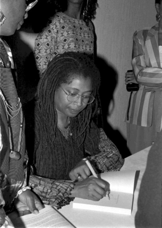 Black woman with long braided hair wearing glasses, sitting and signing a book.