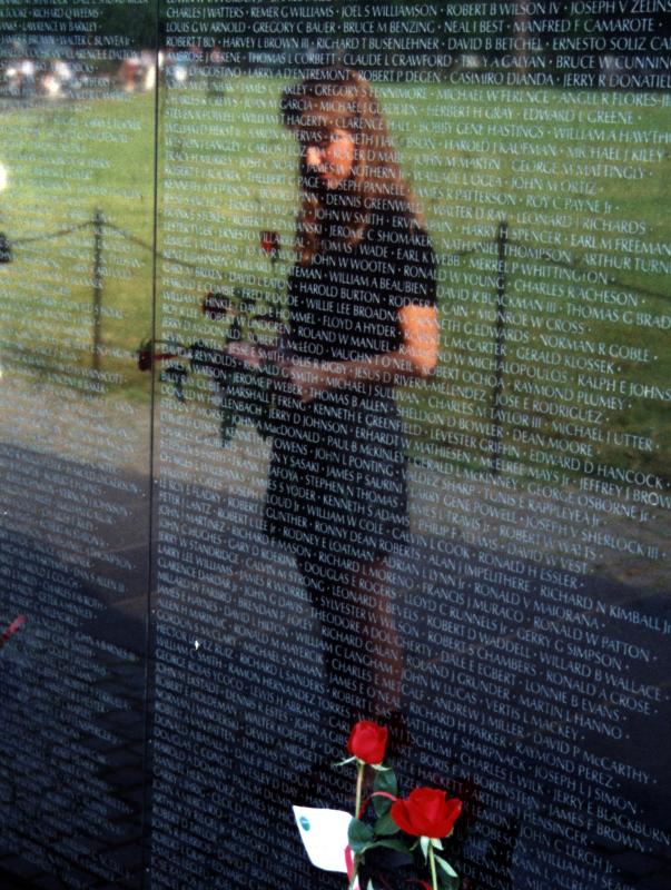 Reflection of a woman in a dress standing in front of a black reflective monument with names engraved on it, a flower at her feet.
