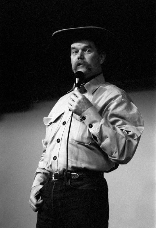 Man with moustache wearing a cowboy hat and white shirt holding a mic on stage. 