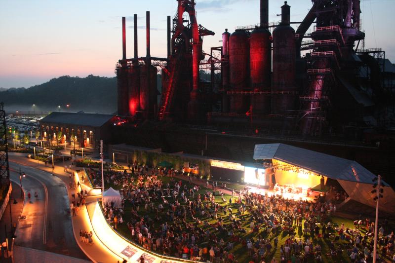 A performing arts venue full of audience members in front of an abandoned steel factory. 
