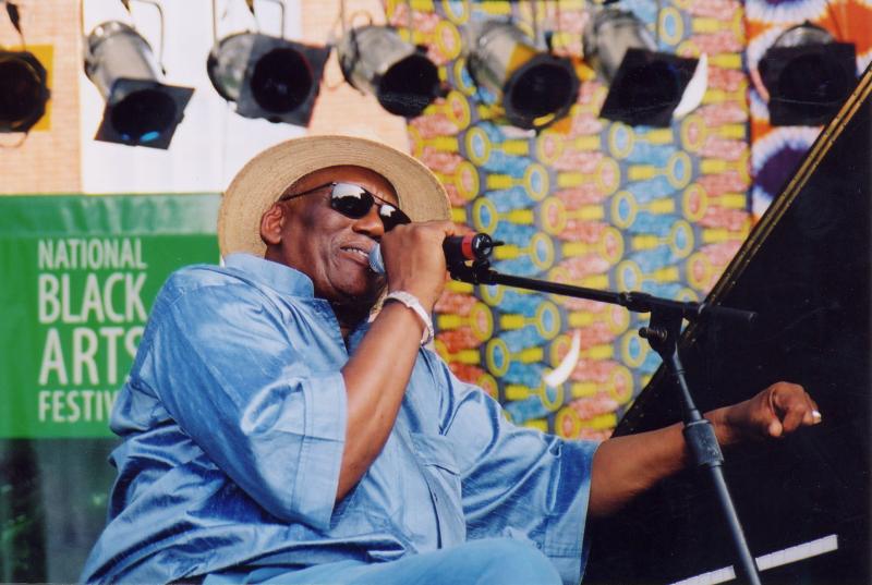 Black man wearing straw hat and sunglasses and blue shirt at piano speaking into a mic on an outdoors stage. 