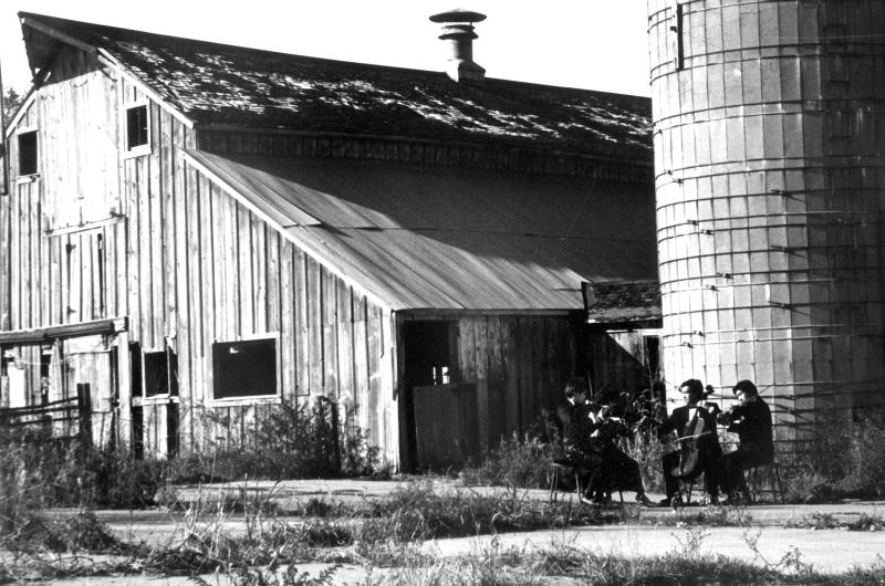 A string quartet playing in front of a barn and silo outdoors.