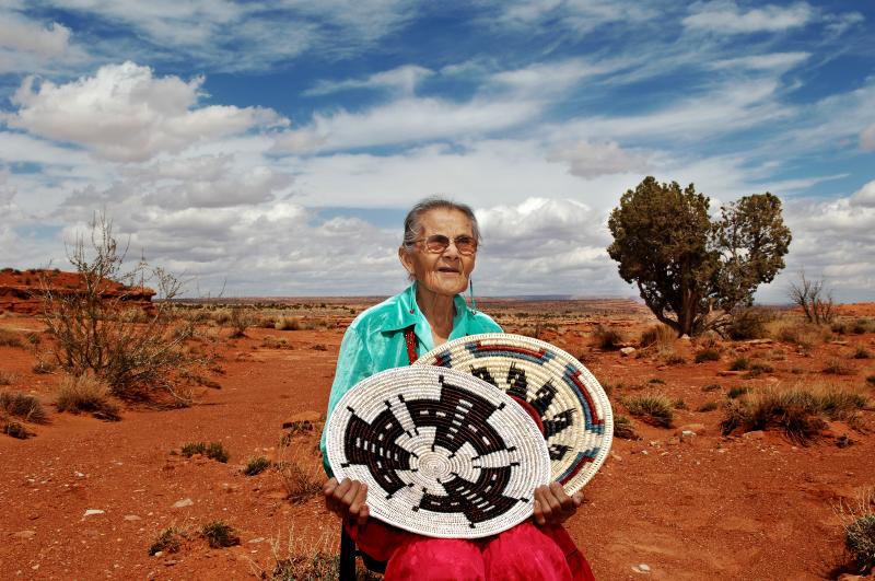 An elderly woman with glasses sits in the high desert holding two handwoven Navajo baskets. The red clay soil stands against the stark landscape and bright sky streaked with clouds. 