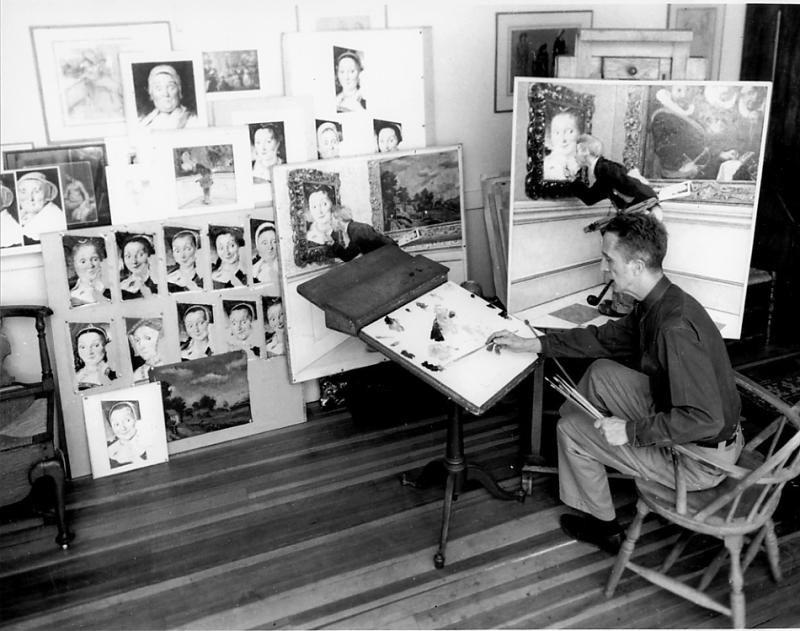 White man smoking a pipe sitting in a wood chair painting a picture in a studio with artwork leaning against a wall. 