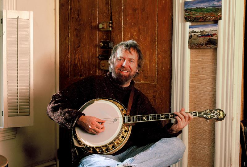 Mick Moloney Portrait by Tom Pich A man with brown hair and a beard smiles while strumming his tenor banjo
