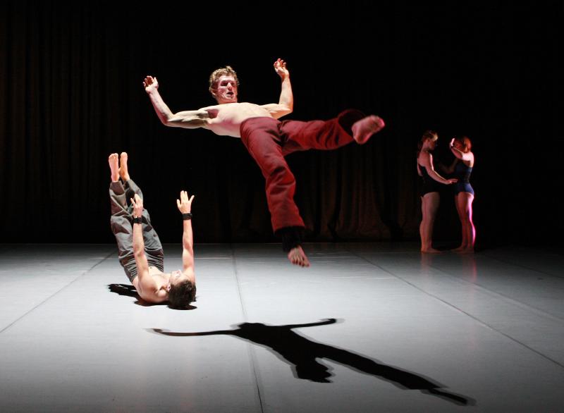 A shirtless white man wearing red pants and barefoot is jumping in the air while another shirtless man is on the ground behind him, and two women to the right, as part of a dance performance. 