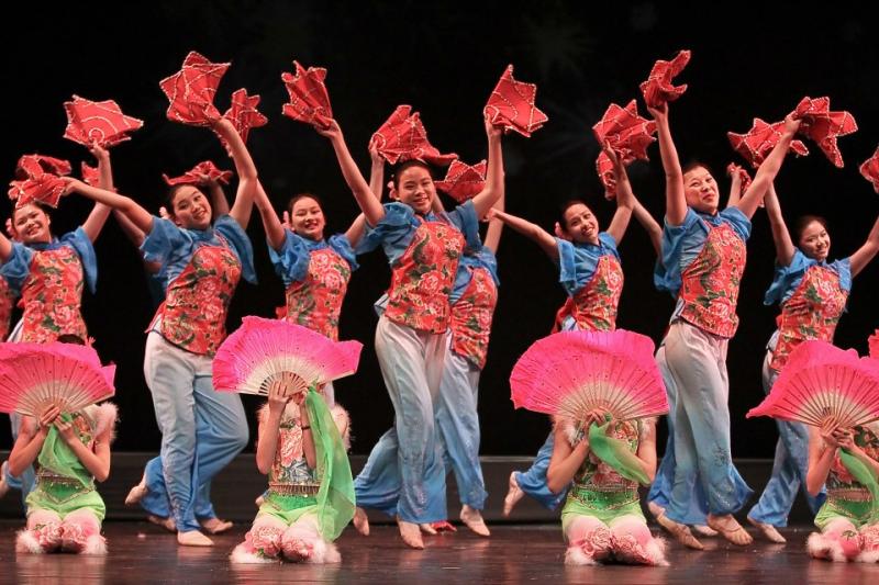 Chinese American girls dressed in blue and pink holding fans and dancing on stage. 
