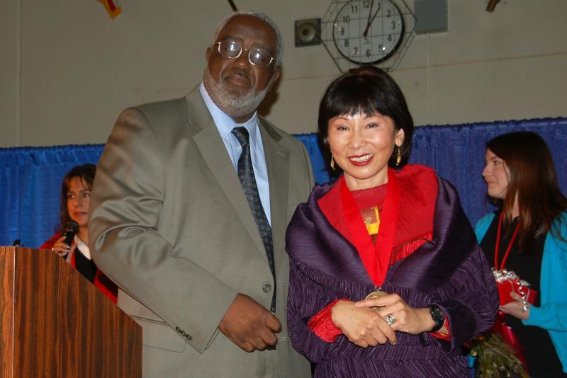 A Black man with a gray beard wearing a gray suit posing next to an Asian woman with dark hair wearing a purple dress. 