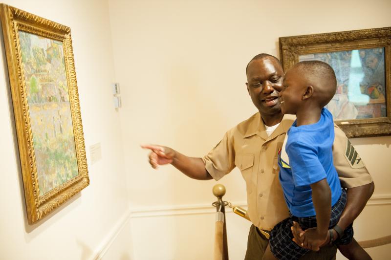 A Black man in a military uniform holds his son up to look at a piece of art in a museum. 