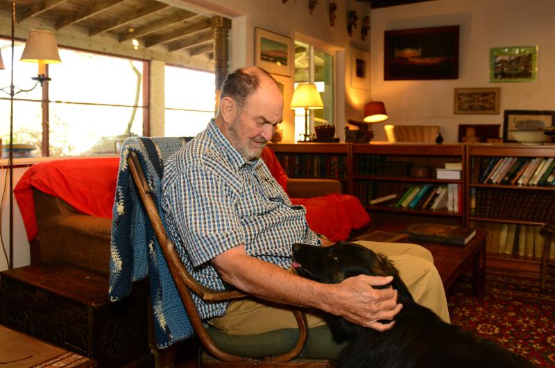 A man sits in a wooden rocking chair petting a black dog with bookshelves and large windows in the background 