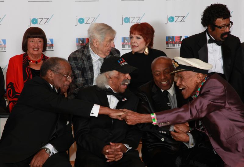 A group of men and women sitting in chairs, two men in the front shaking hands, waiting to pose for a portrait. 