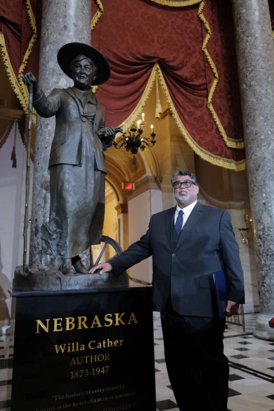 Black man in black suit standing by his bronze sculpture of Willa Cather (who is holding a book in her left hand and a walking stick in her right hand, with grasses and a wheelbarrow near her feet). Black man in black suit standing by his bronze sculpture of Willa Cather (who is holding a book in her left hand and a walking stick in her right hand, with grasses and a wheelbarrow near her feet).