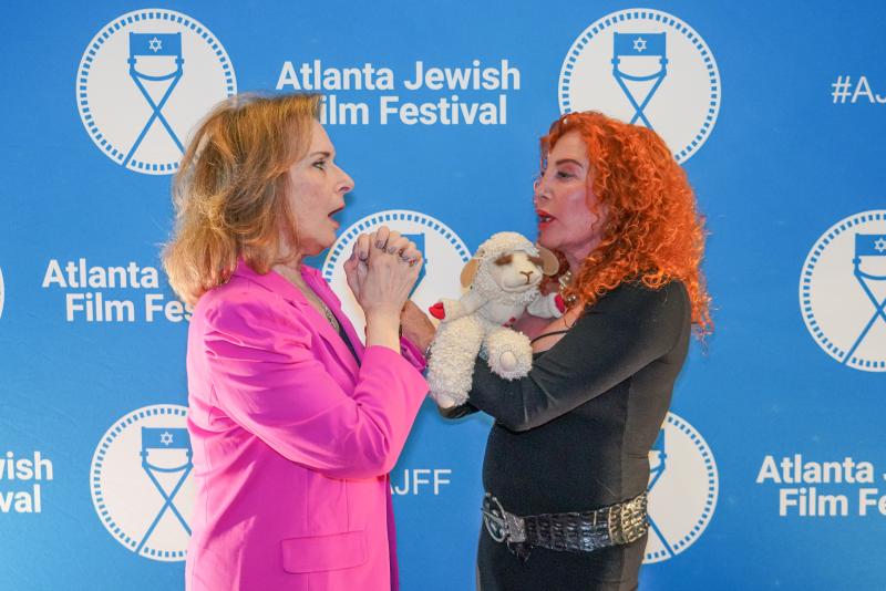 Photo of two White women: woman on left has dirty blonde hair and is wearing a pink blazer and the woman on the right has red curly hair and is wearing a black outfit and holding a stuffed lamb toy. Behind the women is a blue and white step and repeat with “Atlanta Jewish Film Festival” written on it.