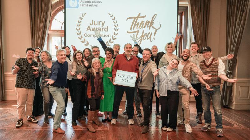 Photo of a group of people holding their arms out in celebration and smiling in front of a projector that says “Atlanta Jewish Festival,” “Jury Award Competition,” and “Thank You!”