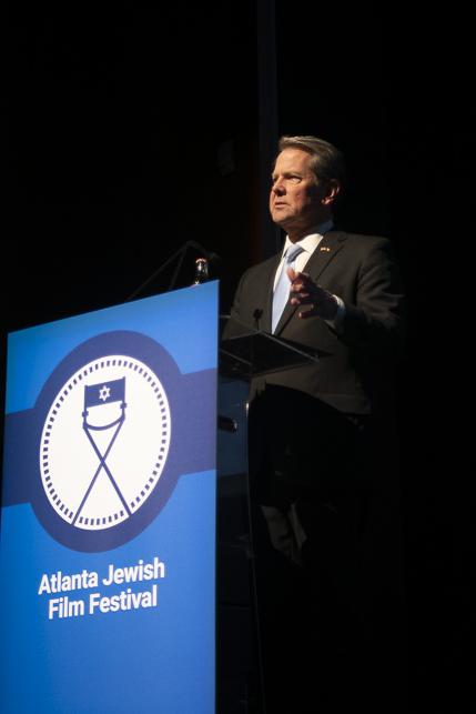 Photo of a white man in a black blazer, holding a mic and standing at a podium. The podium (blue and white colors) has “Atlanta Jewish Film Festival” written on it.