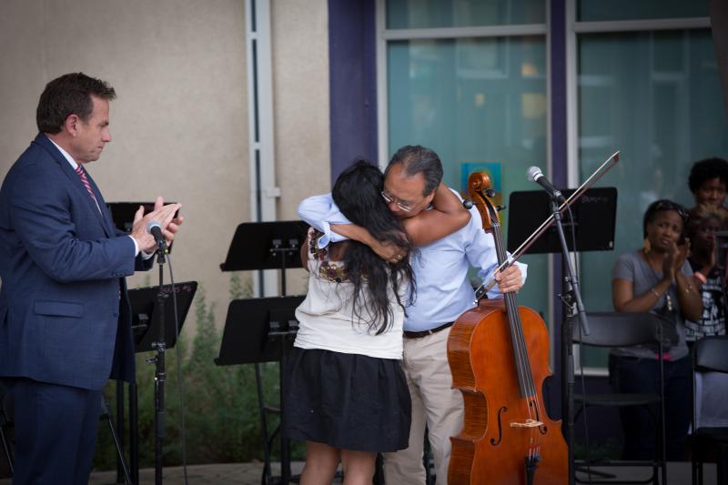 A White man in a blue suit (left) claps both of his hands while facing a woman with long, black hair (center) who is hugging Yo-Yo Ma, an Asian man with a blue button-down shirt and khaki pants.