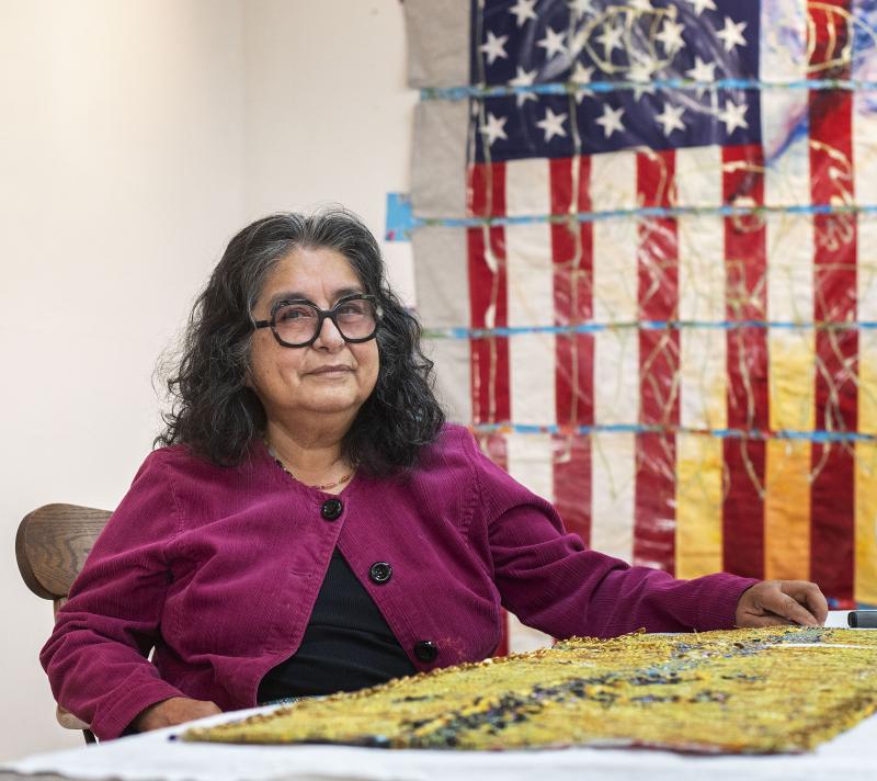 Hispanic/Latinx woman wearing black glasses and a purple cardigan sitting at a table with her artwork. An artwork featuring the American flag is behind her on the right. 