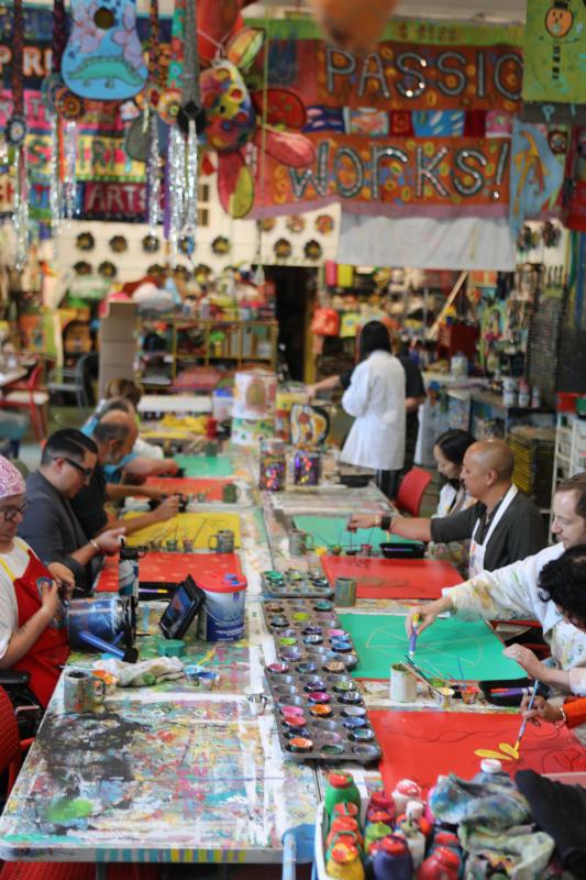 Alt text: People sit at a long table with colorful red and green sheets in front of them that they are painting. Above them hang colorful artwork including a banner reading “Passion Works!”