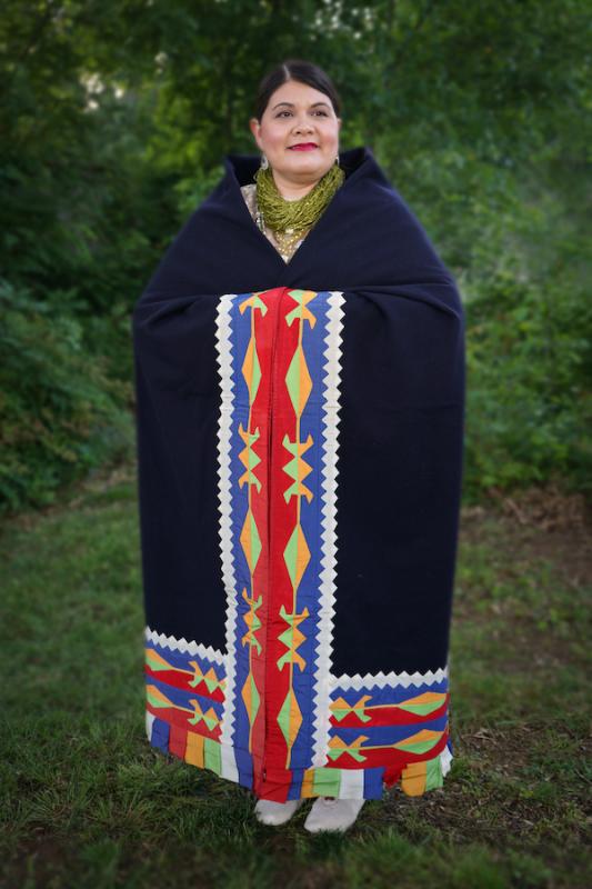 A woman with dark hair wears a long, dark blue blanket with colorful ribbonwork stitched into the border in a blue, red, green, and yellow pattern with angular white edge.