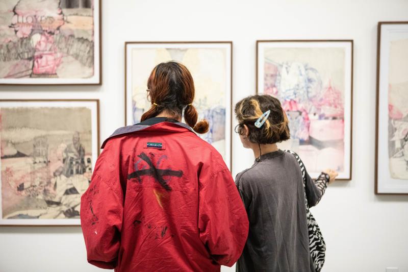 Two people with their backs to the viewer stand looking at a series of art works hung on a wall in a gallery