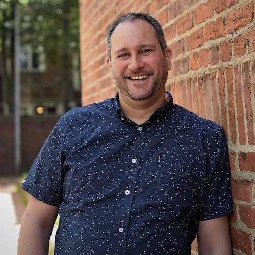 Aaron Kubey is a white man in his forties with a fair skin complexion, gray hair and beard. Aaron is wearing a dark button-down shirt with short sleeves, white buttons and colorful dots all over it. He is smiling and leaning against a brick building wall.